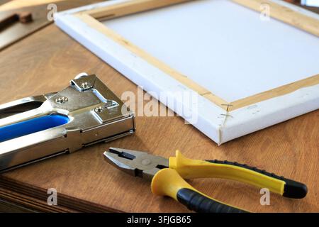 Canvas stretching tools with a metal staple gun and pliers placed next to a wooden frame on a wooden surface in an art workshop. Stock Photo