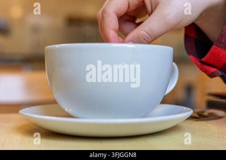 Close-up of fingers dropping something into large white ceramic coffee cup on matching saucer, person wearing red plaid flannel shirt, warm cafe-style Stock Photo