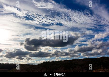 Dramatic sunlit clouds drifting across a vibrant blue sky over a dark rugged hill, creating a stunning contrast between light and shadows in this capt Stock Photo