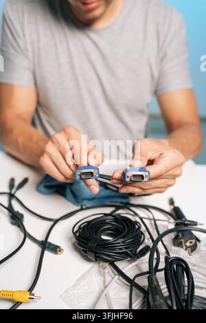 Vertical cropped shot of man holding two VGA connectors, preparing collection of cables, cords, electronic accessories on table. Concept of process of Stock Photo