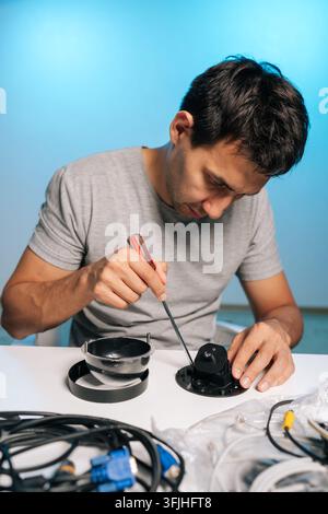 Vertical portrait of technician using screwdriver to assemble, repair dome security camera, working on components, wiring on white table against blue Stock Photo