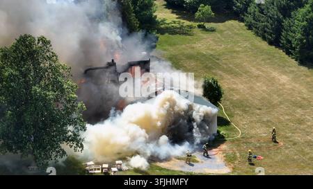Firemen in protection clothes spraying water with hose on burning house in rural area. Sunny day on farmland of american town. Dense toxic fumes risin Stock Photo
