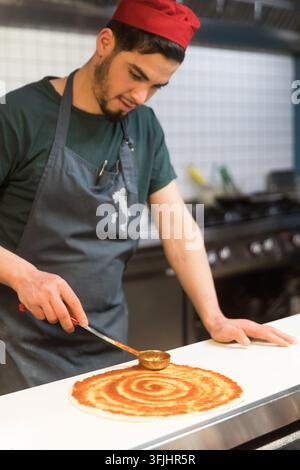 A chef in an Italian pizzeria uses a ladle to spread tomato sauce onto dough, preparing a pizza for baking. Stock Photo