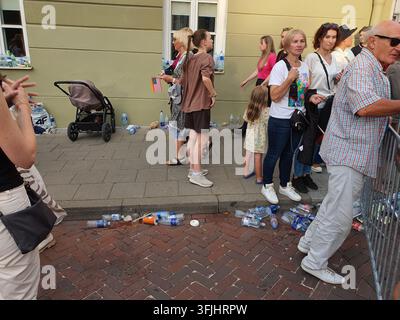 A group of people standing in a queue on a sidewalk next to a pale yellow building with white window frames. Several discarded plastic water bottles Stock Photo