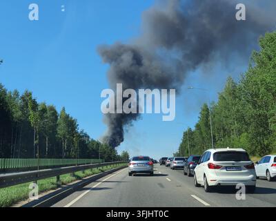 Thick black smoke billows into a clear blue sky from a vehicle fire on a busy multi-lane highway, causing a traffic jam with numerous cars stopped on Stock Photo