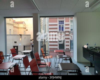 An empty modern classroom or lecture hall with red chairs and dark tablet desks arranged in rows. Large floor-to-ceiling windows reveal an urban Stock Photo