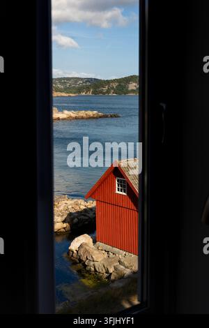 View from a window at Lindesnes on the southern coast of Norway, overlooking the sea, rocky shoreline and a traditional red wooden boathouse. Stock Photo