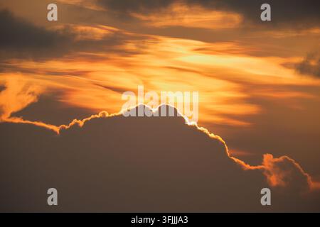 Vibrant golden and orange sunset sky illuminated behind a large, dark cloud formation with a glowing silver lining during dusk. Stock Photo