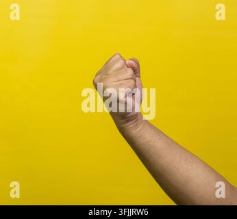 A right arm is raised, forming a tightly clenched fist held upward against a solid, bright yellow background. The knuckles are pronounced, and the fin Stock Photo