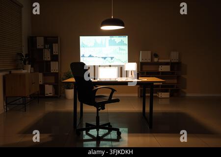Interior of data scientists office with workplace, shelving units, documents, laptop and diagrams projection on wall in evening Stock Photo
