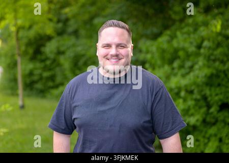 Friendly young man smiling naturally at the camera while standing outdoors in a lush green setting, expressing confidence, approachability, happiness Stock Photo