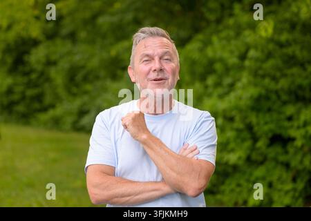 Relaxed mature man smiling confidently with folded arms while standing outdoors in a natural green environment, expressing experience, trust, optimism Stock Photo