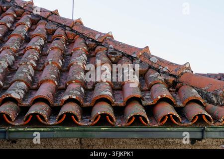 Old terracotta roof tiles covered with lichen above a green metal gutter and stone wall. Traditional clay roofing for construction, renovation and Stock Photo