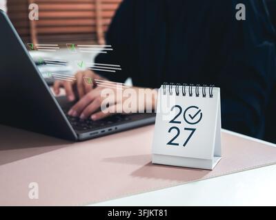 Desk calendar marked 2027 beside businessperson typing on laptop with digital checklist icons. Annual business planning, goal achievement, task manage Stock Photo