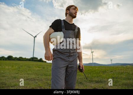 Wind turbine technician holding hard hat and radio in a field at sunset Stock Photo