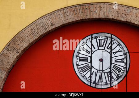 A large clock painted on a vibrant, flat red wall framed by a classic brick archway in Perugia, Italy. Stock Photo
