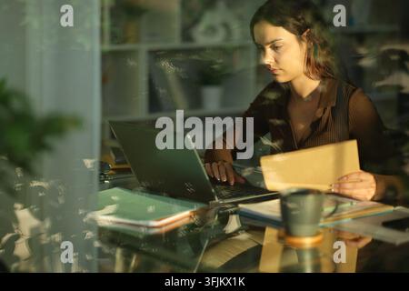 Concentrated telecommuter working online holding documents and checking laptop at home Stock Photo