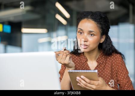 Businesswoman working on laptop and taking notes in office, professional woman using computer for financial analysis and planning at her desk in workplace. Focused employee reviewing documents. Stock Photo