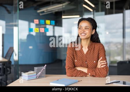 Career success and professional growth, smiling businesswoman sitting at desk in modern office with sticky notes on glass wall for project planning. Confident employee working on strategy and goals. Stock Photo