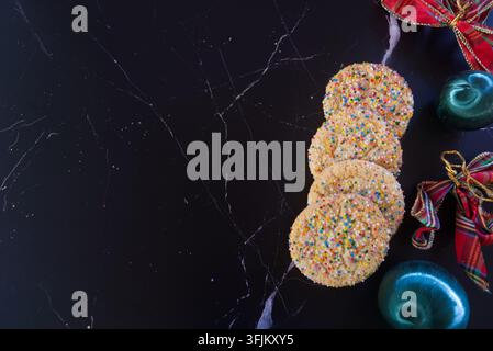 Colorful sprinkle-covered Christmas cookies arranged with festive ornaments and plaid ribbons on a dark countertop, creating a cheerful holiday food c Stock Photo