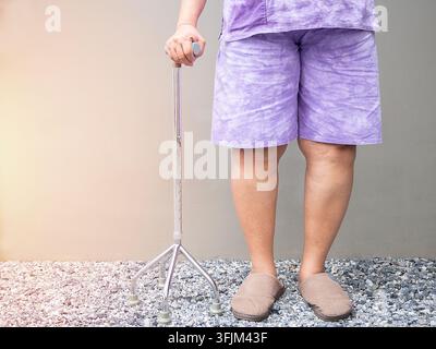 Cropped image of Asian senior woman standing and using a walking stick at home with light gray background. Stock Photo