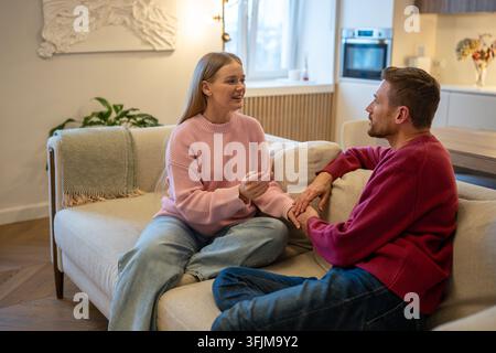 Positive woman speaking lively to loving man partner while married couple sitting on couch at home. Stock Photo