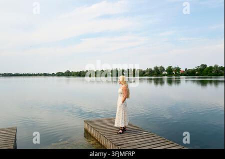 Blonde woman in sundress looks over peaceful lake Stock Photo