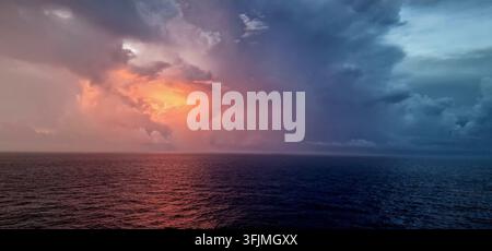 Dramatic rainstorm over the open ocean illuminated by orange sunset light beneath dark storm clouds. Stock Photo
