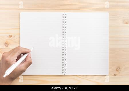 Top view of hand holding white pen writing on open lined spiral notebook mockup on light wooden background with copy space Stock Photo