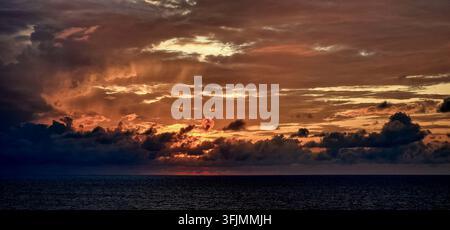 Dramatic red and orange sunset illuminating layered clouds over the dark open ocean. Stock Photo