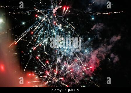 Multicolored fireworks burst and streak across a dark night sky, trailing smoke and glowing red and white sparks in overlapping starburst patterns. Stock Photo