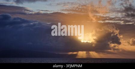 Dramatic sunlight breaking through dark storm clouds over the open ocean, creating golden sun rays above the horizon. Stock Photo