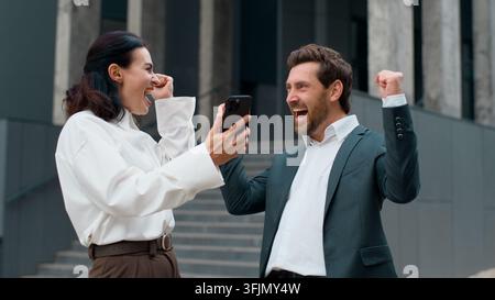 Two happy Caucasian middle-aged business colleagues winning online with mobile phone in city. Positive man with woman businesswoman sharing good news Stock Photo
