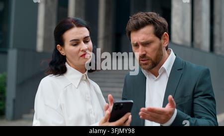 Two Caucasian business workers looking in mobile phone upset with reading bad news in city. European middle-aged man with woman sad with smartphone Stock Photo