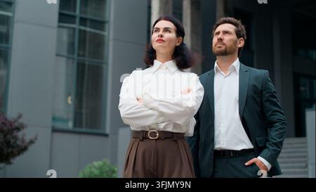 Two confident middle-aged Caucasian business people crossing hands looking away thinking dreamy in city. Mature man woman formal businessman with Stock Photo
