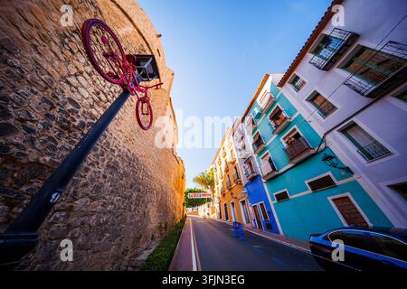 Wide perspective along a street in Villajoyosa, showing a red bicycle lamp post next to the old stone wall on one side and brightly painted blue and t Stock Photo