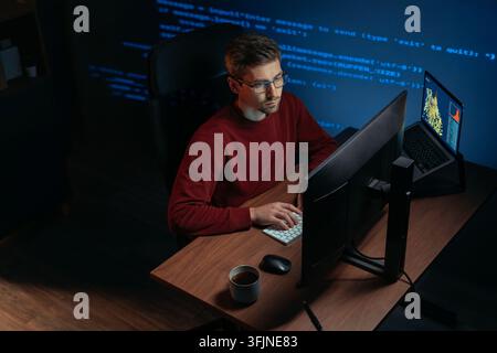 Software engineer wearing eyeglasses working at multi-screen desk in a dark office, focused on code and graphs. Programmer works late at night on a demanding data science and AI task. Copy space Stock Photo