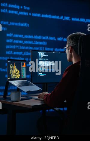 Focused coder works at desk in a dark office with programming code lines running behind. Developer is deeply engaged in a late night data science or AI coding session. Copy space.  Stock Photo