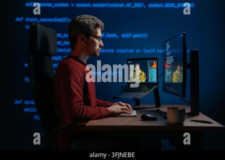 Side view, coder at a dark office desk works across multiple screens at night, with programming script on a background. Data scientist working on a machine learning or artificial intelligence task. Stock Photo