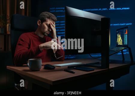 Exhausted software engineer working overtime in a dark home office, taking off glasses and rubbing tired eyes after hours of coding in front of screen and under deadline pressure.  Stock Photo