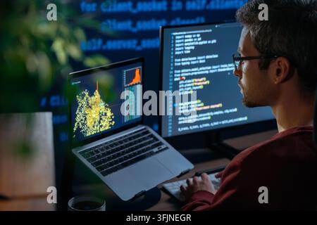 Over shoulder view, data scientist in glasses sits at a dark desk, surrounded by multiple monitors displaying programming scripts and graphs, working late at night on artificial intelligence project Stock Photo