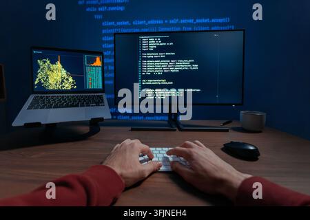 First person view of a programmer hands typing on a keyboard at night, two monitors displaying code and data science visualizations on dark desk, works on a machine learning or data science. POV Stock Photo