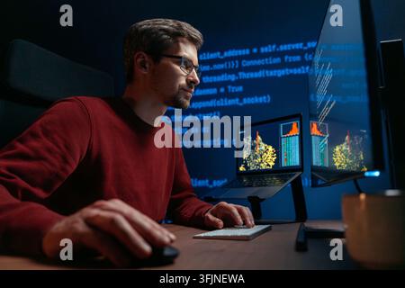 Smiling male coder in glasses works at a dark office desk, surrounded by screens filled with programming code and graph, solving a complex AI or data science tasks late at night. Stock Photo