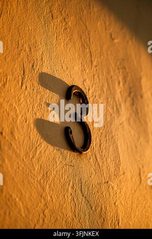 Metal numeral three mounted on a stucco wall bathed in warm sunlight. Concept of residential address identification, architectural detail, house numbe Stock Photo