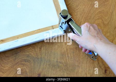 Canvas stretching with a hand holding a chrome staple gun to fix fabric onto a wooden stretcher frame, top view on a wooden table. Stock Photo