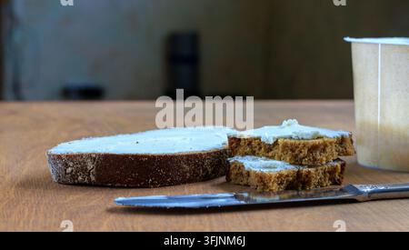 Dark rye bread slices with cream cheese spread placed near a knife and a plastic container on a wooden table, side view. Stock Photo