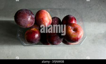 A collection of ripe, colorful plums are neatly arranged in a clear plastic store-bought container on a light, neutral surface. This image is suitable Stock Photo