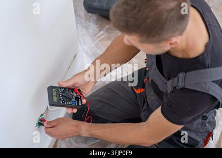 Man tests electrical wires in a wall with a digital multimeter. Electrician checks the wiring before installing an outlet. Home electrical work, repai Stock Photo