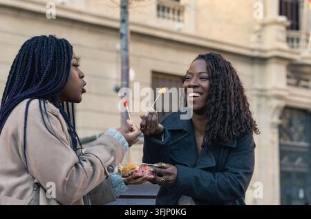 Two cheerful young women enjoying a fruit snack together on a city bench, sharing a moment of happiness and friendship Stock Photo