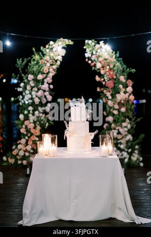 A three-tiered white wedding cake with delicate feather-like decorations is displayed on a white tablecloth, surrounded by glass candle holders with l Stock Photo